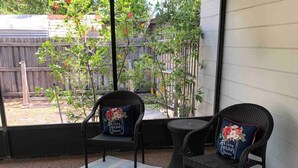 A covered patio area with two wicker chairs, a striped rug, and a view of a backyard with a wooden fence and greenery.