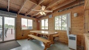 Una cabaña de madera con una mesa grande, bancos, un ventilador de techo y un calentador.