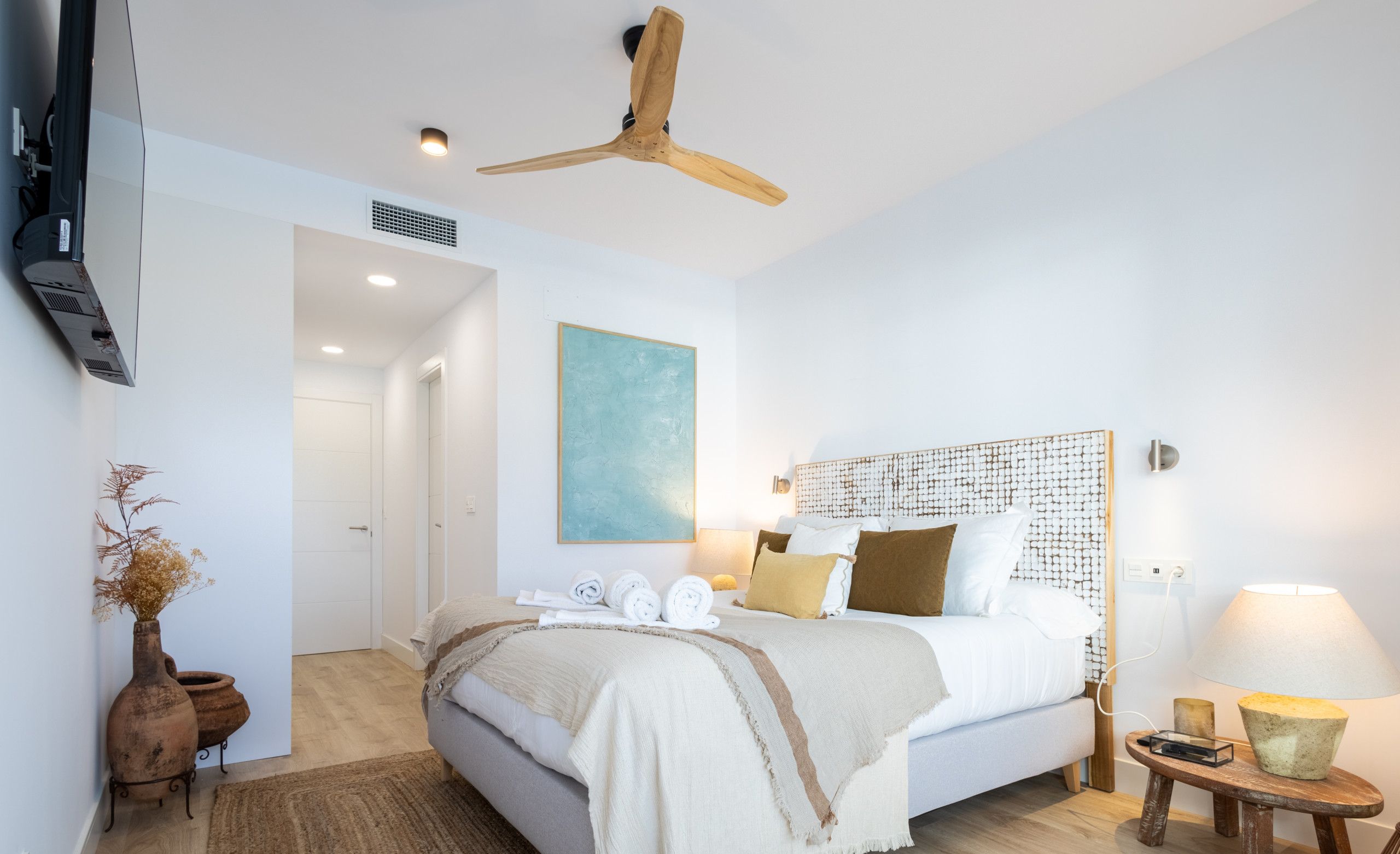 Een moderne slaapkamer met een groot bed, een plafondventilator, een schilderij aan de muur en een nachtkastje met een lamp.