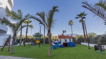 Un parque infantil con columpios, toboganes y estructuras para escalar, rodeado de palmeras y un cielo despejado.