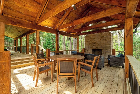 Un espacio al aire libre techado con chimenea de piedra, mobiliario de madera y vista al bosque.