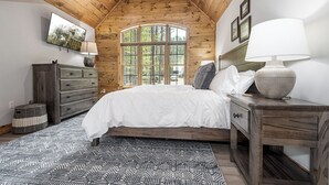 Un dormitorio con techo de madera, un ventanal grande, cama con ropa de cama blanca, cómoda, mesita de noche con lámpara y alfombra estampada.