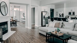 A modern living room with a fireplace, a sofa, and a coffee table. There is a kitchen area with white cabinets and stainless steel appliances.