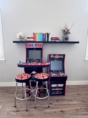 Una habitación con dos máquinas de videojuegos, una estantería con libros y un globo terráqueo, además de dos taburetes.
