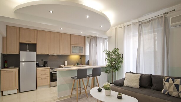 A modern kitchen with wooden cabinets, a built-in oven, and a refrigerator. There is a dining area with a white table and two chairs. A sofa with cushions is in the living area, and a plant is on the table.
