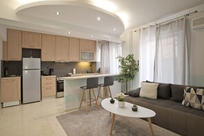 Una cocina moderna con armarios de madera, horno empotrado y refrigerador. Hay un comedor con mesa blanca y dos sillas. Un sofá con cojines se encuentra en la sala de estar, y una planta está sobre la mesa.