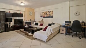 A living room with a sofa, coffee table, TV, fireplace, and a desk with a computer. - Paradeisos Modern, 2BR, 2BA, Fully Equipped (Chalandri)