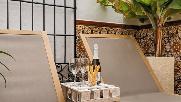 Une chambre avec un lit, une petite table sur laquelle se trouvent une bouteille et des verres, une plante et une fenêtre donnant sur un paysage.