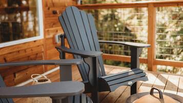 A wooden deck with Adirondack chairs and a table, a metal lantern, and a large window overlooking greenery.