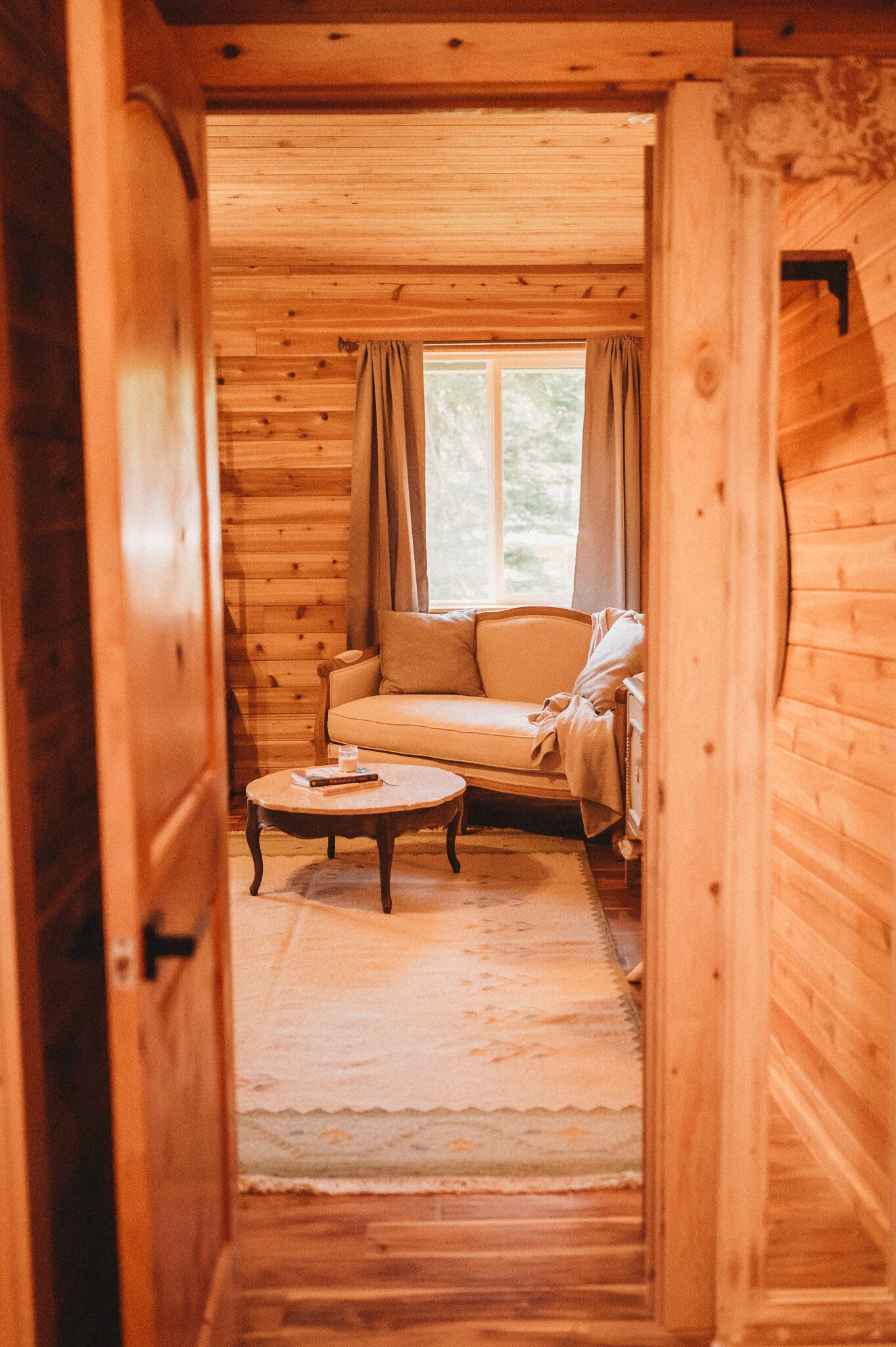 Un interior de madera con un sofá, una mesa de centro redonda y una ventana con cortinas.