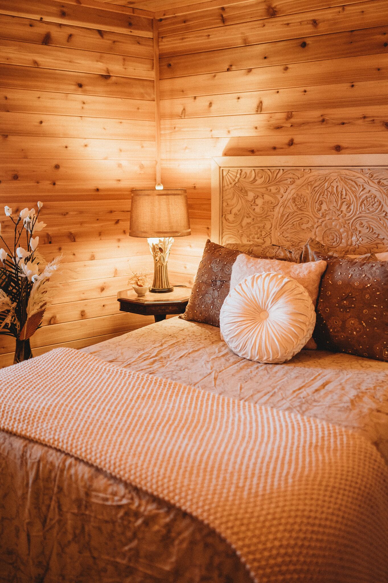 Un dormitorio acogedor tipo cabaña de madera, con cama, almohadas, mesita de noche, lámpara y un florero con flores.