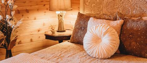 Un dormitorio acogedor tipo cabaña de madera, con cama, almohadas, mesita de noche, lámpara y un florero con flores.
