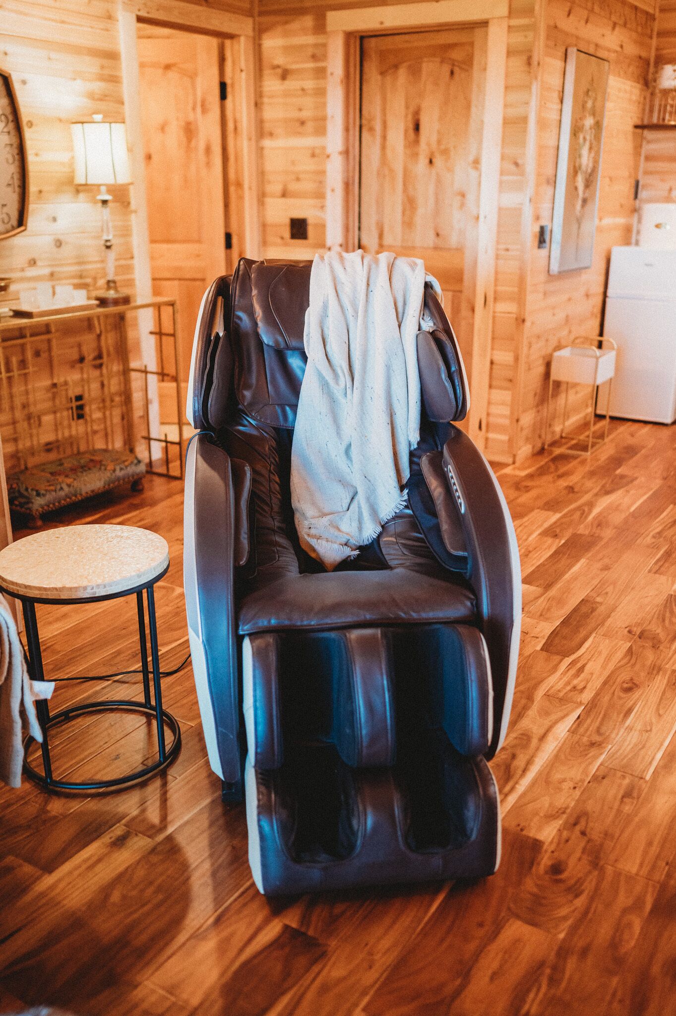 A massage chair in a wooden cabin room with a small round table and a lamp.