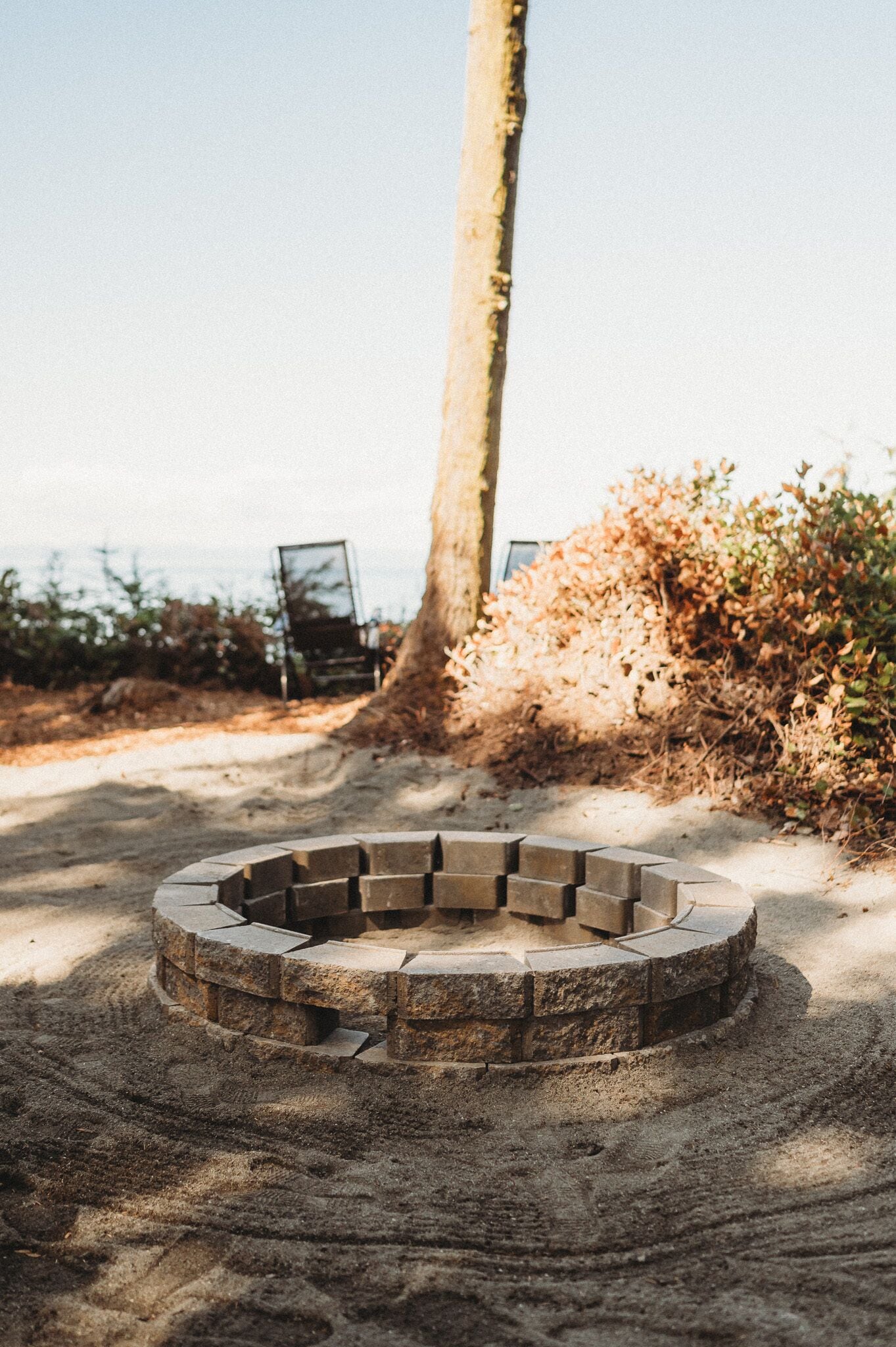 Un hogar circular de piedra rodeado de arena, con un árbol y vegetación seca al fondo.