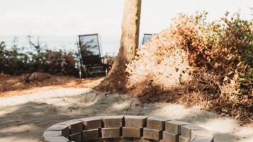A circular stone fire pit surrounded by sand, with a tree and dry foliage in the background.