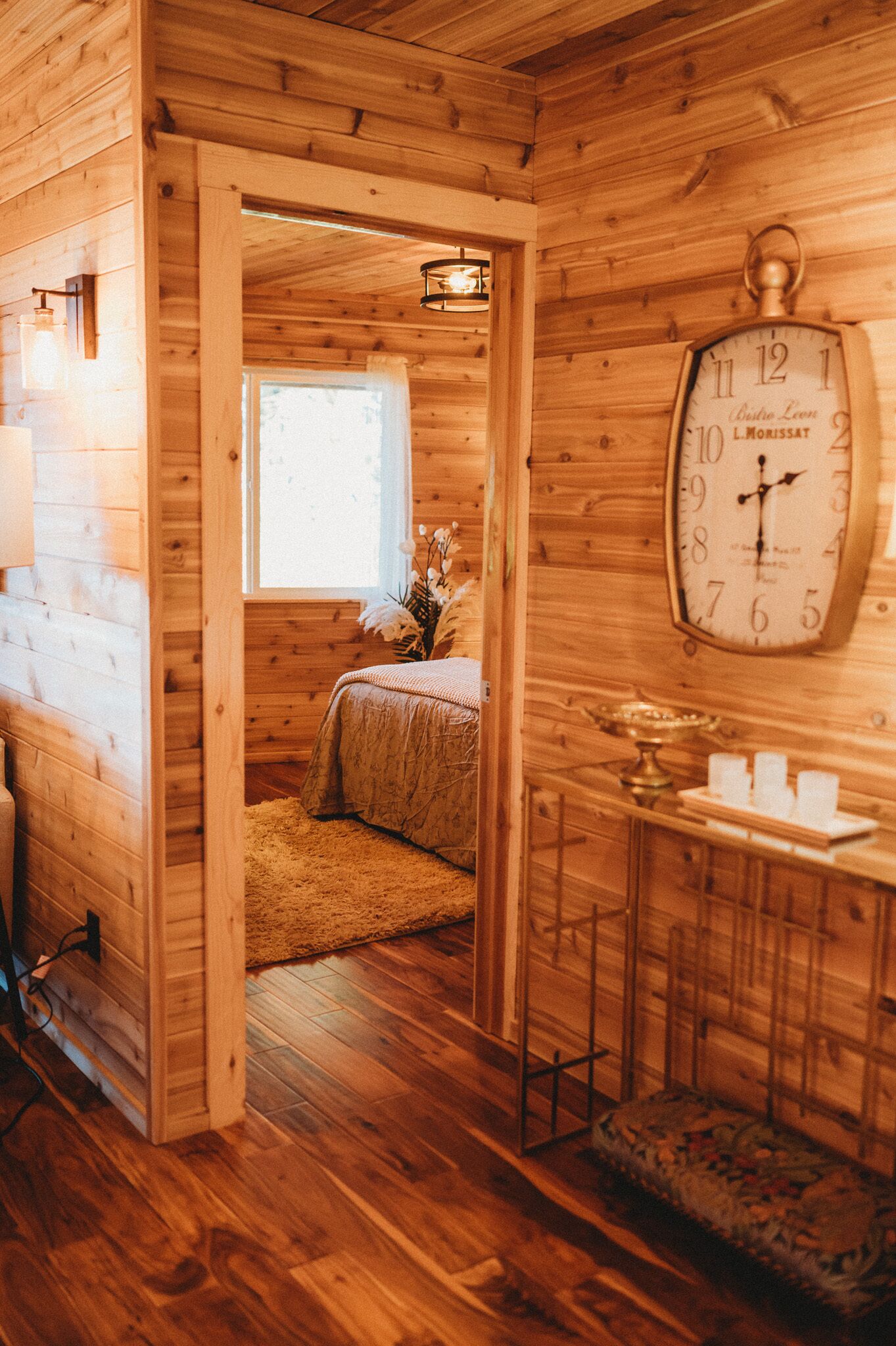 Interior de una cabaña de madera con una cama, un reloj de pared grande y una ventana.