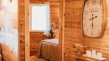 Interior de una cabaña de madera con una cama, un reloj de pared grande y una ventana.
