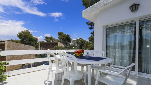 A patio with a round table and white plastic chairs, a bowl of fruit, and a view of the surrounding area.