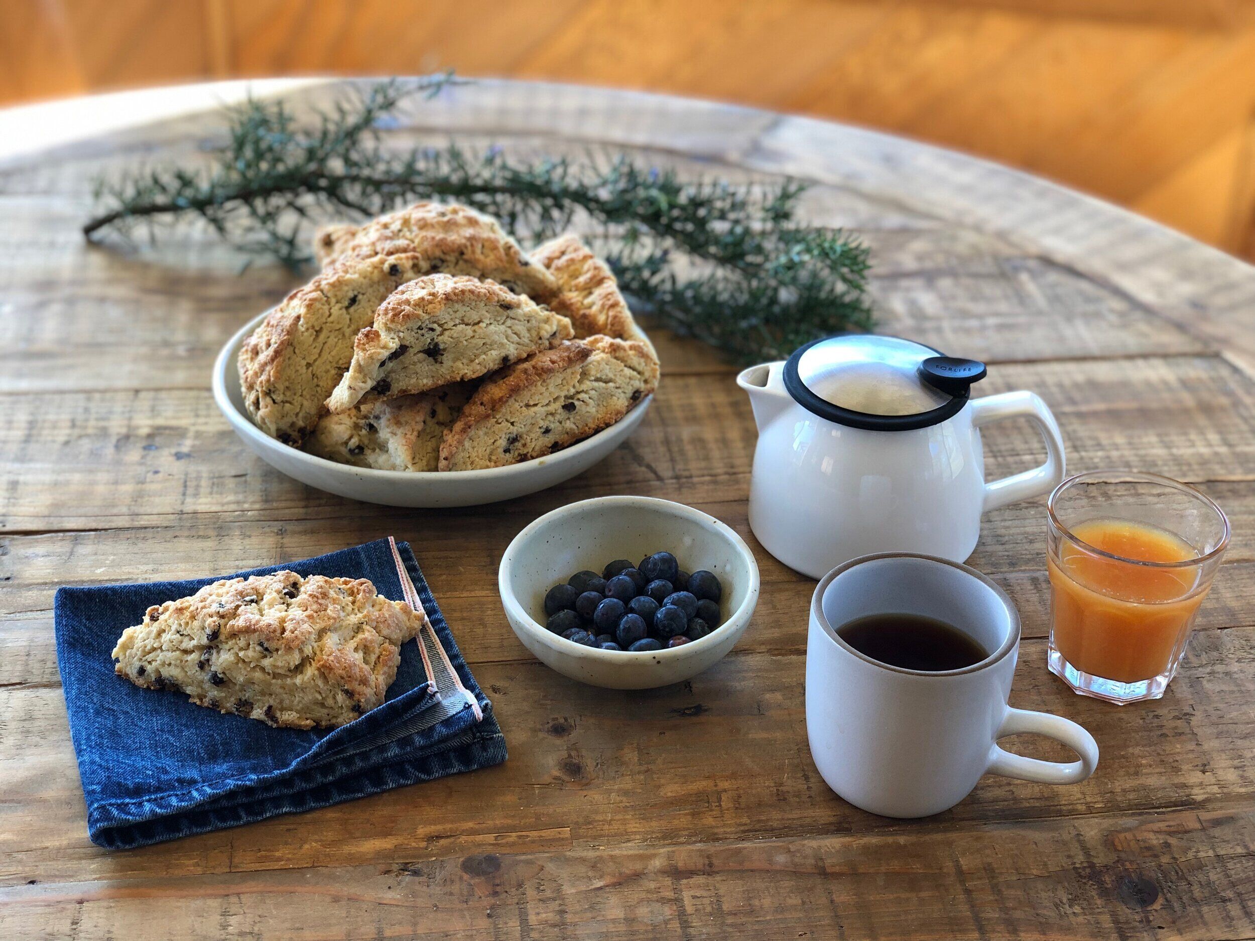 A rustic wooden table with a bowl of scones, a small bowl of blueberries, a coffee pot, a mug of coffee, and a glass of orange juice.
