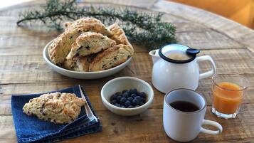 A rustic wooden table with a bowl of scones, a small bowl of blueberries, a coffee pot, a mug of coffee, and a glass of orange juice.