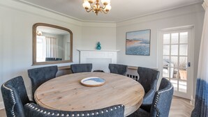 A dining room with a round wooden table, six upholstered chairs with decorative studs, a chandelier, a large mirror, a fireplace, and a painting on the wall. - Downland House - Four Bedroom House, Sleeps 10 (Woolacombe)