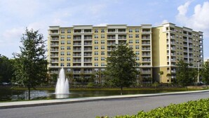 Un hotel de varios pisos con una fuente en primer plano, rodeado de vegetación y un cielo despejado.