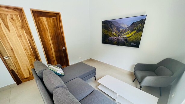 A modern living room with a grey sofa, a white coffee table, and a grey armchair. There is a wooden door and a wall-mounted TV displaying a mountainous landscape. - Villa Estrella Pavones (Pavón)