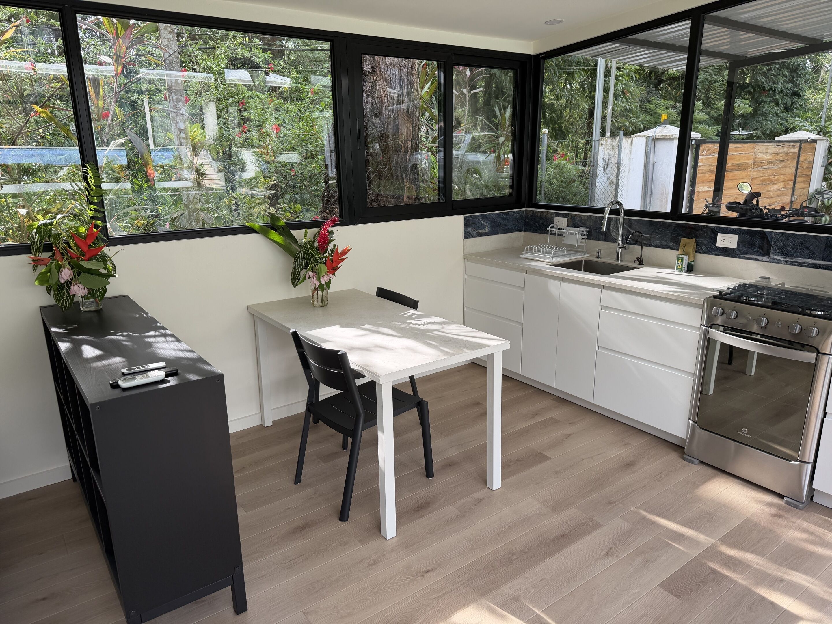 A modern kitchen with a dining table, black chairs, a stove, and a view of the garden through large windows.