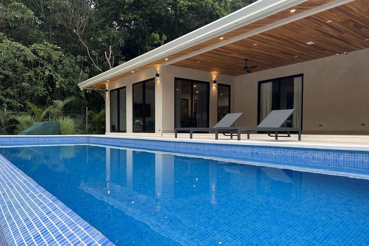 Et moderne poolområde med et klart, blåt pool, liggestole og en overdækket terrasse med store vinduer.