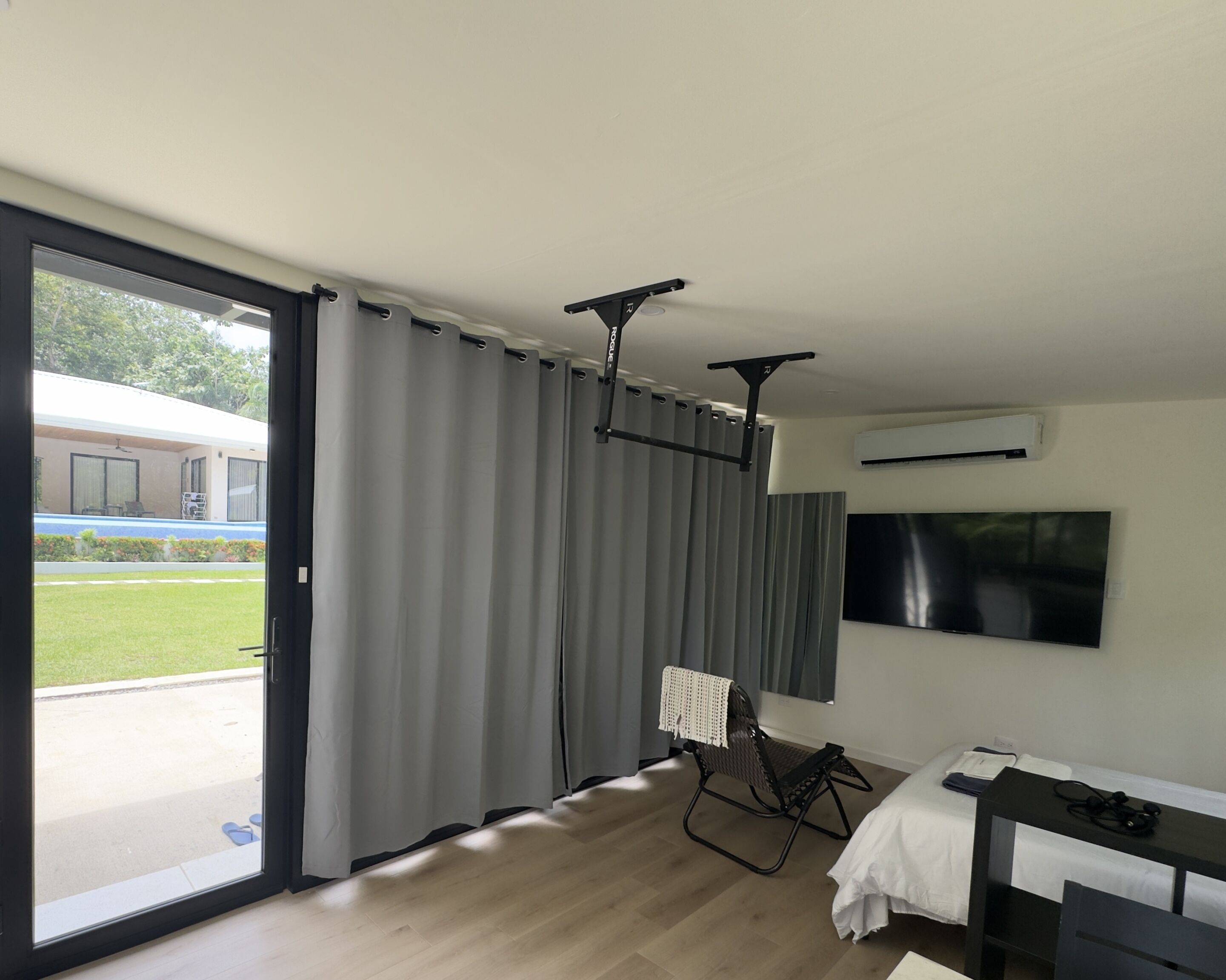 A room with a sliding glass door, a ceiling-mounted air conditioning unit, a mounted flat-screen TV, and a bed with a white bedspread.
