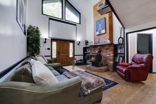 A cozy living room with a fireplace, a red leather chair, a green sofa, and a large window. - SantiCabin – Modern Pocono Mountains Retreat (Albrightsville)