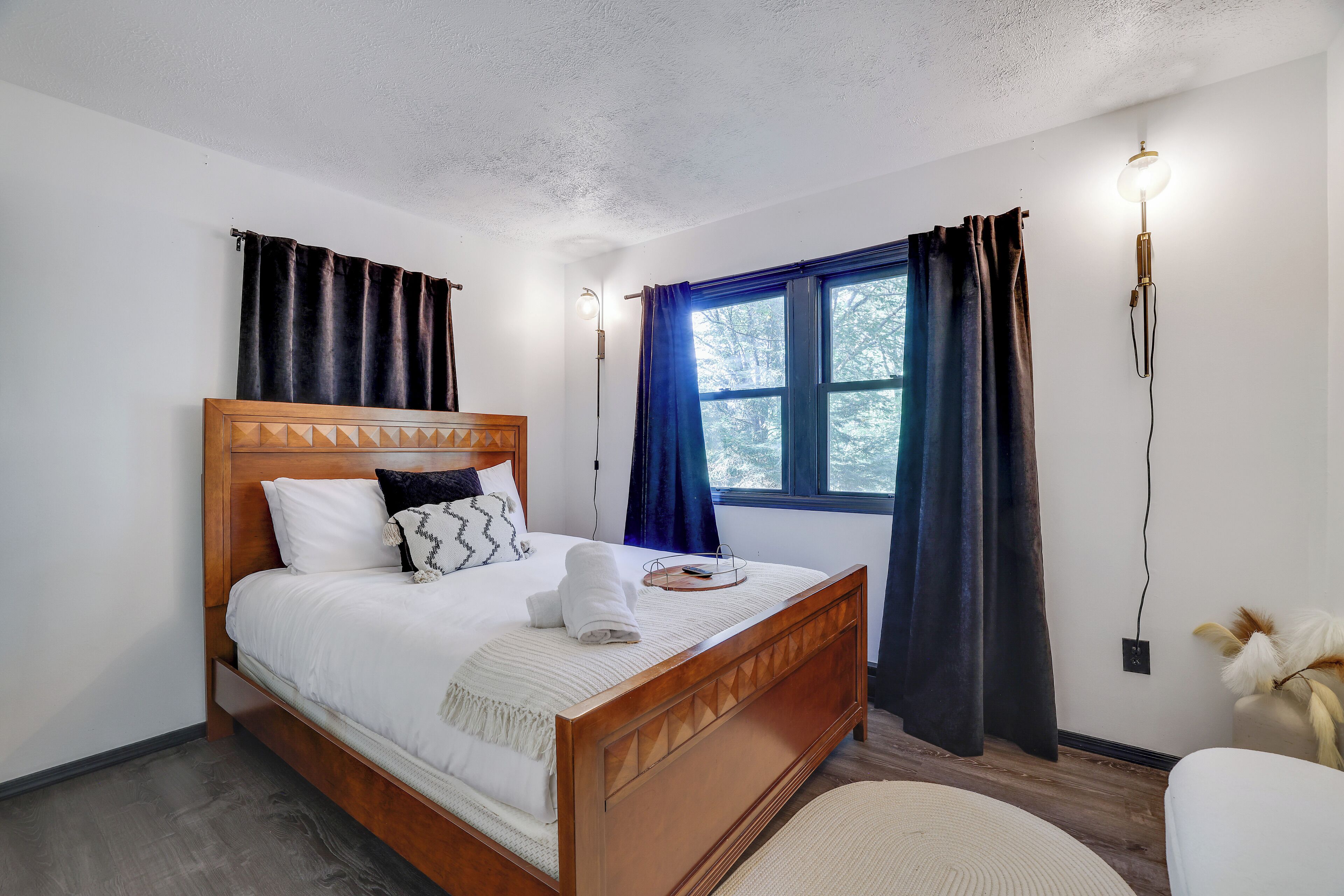 Una cama bien tendida con cabecera de madera, una mesita de noche con lámpara y una ventana con cortinas.