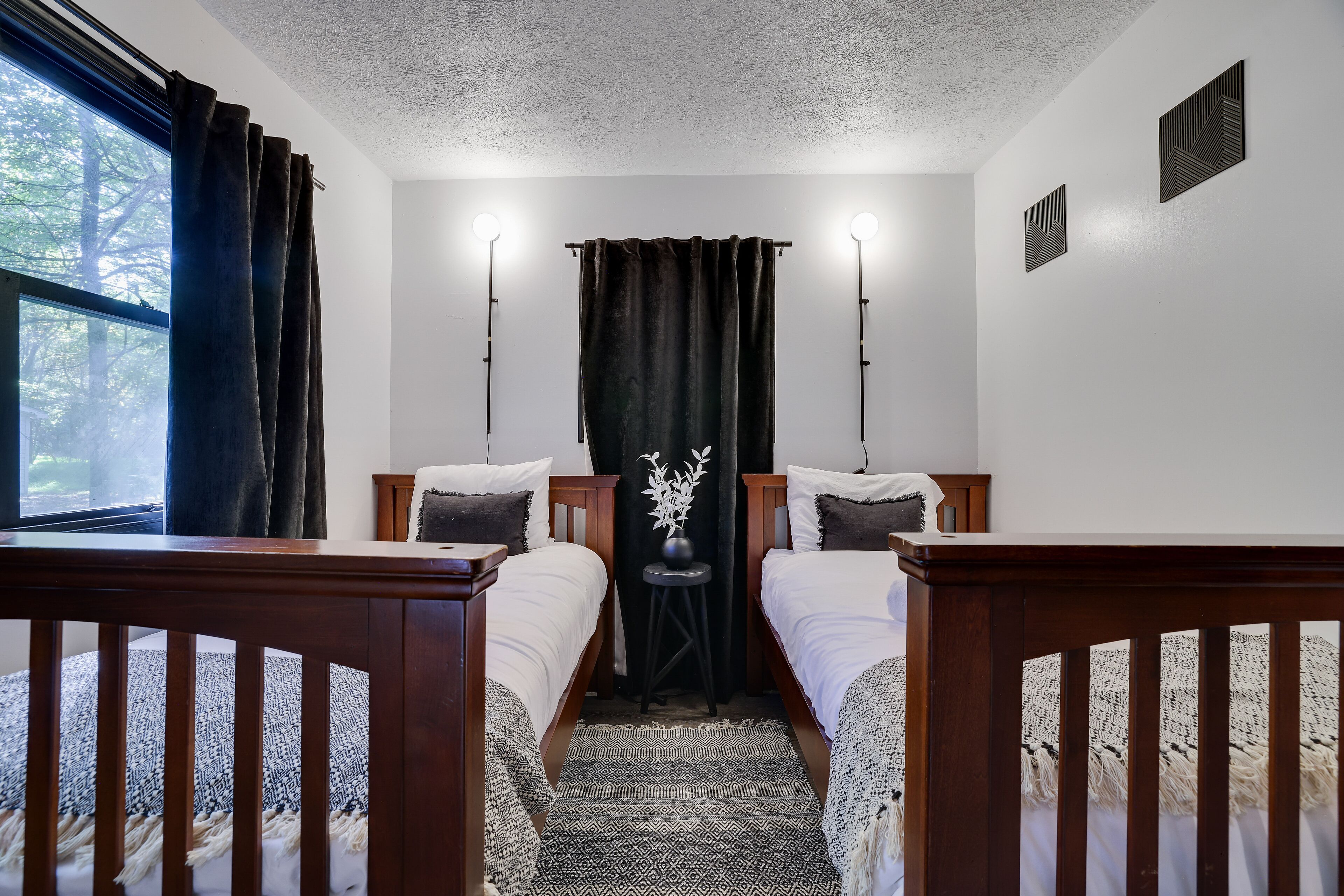 A room with two beds, a window with curtains, a small table with a vase, and wall-mounted lights.