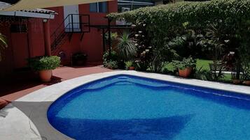 Una piscina con un edificio rojo al fondo, rodeada de plantas en macetas y una escalera.