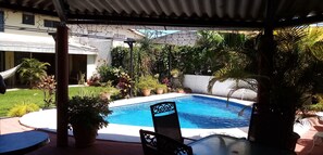 A swimming pool with a chair and a sign, surrounded by potted plants and a covered area. - Casa San Benito (San Salvador)