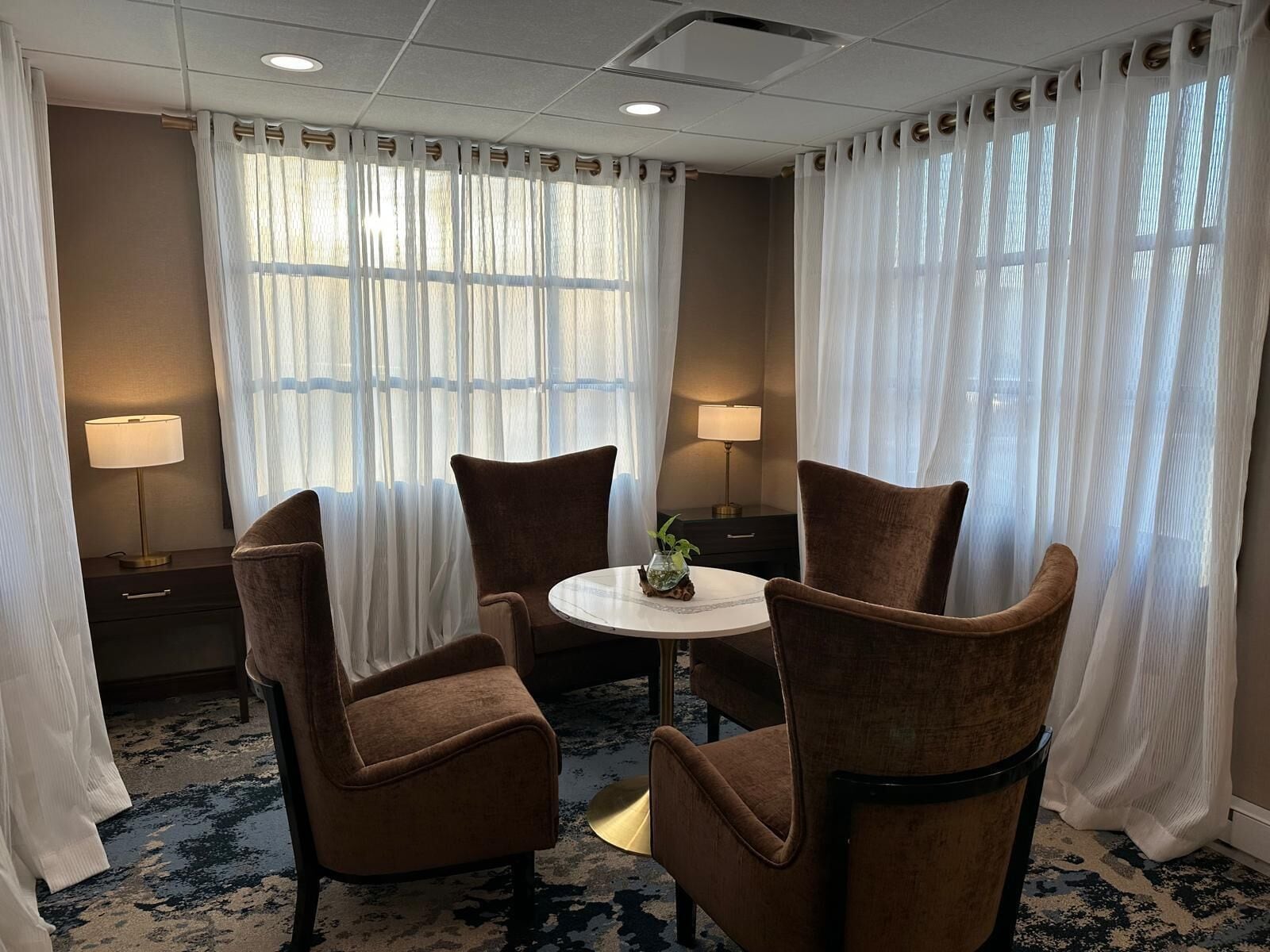 Une chambre d’hôtel avec une table ronde, deux chaises marron, une petite plante et deux lampes.