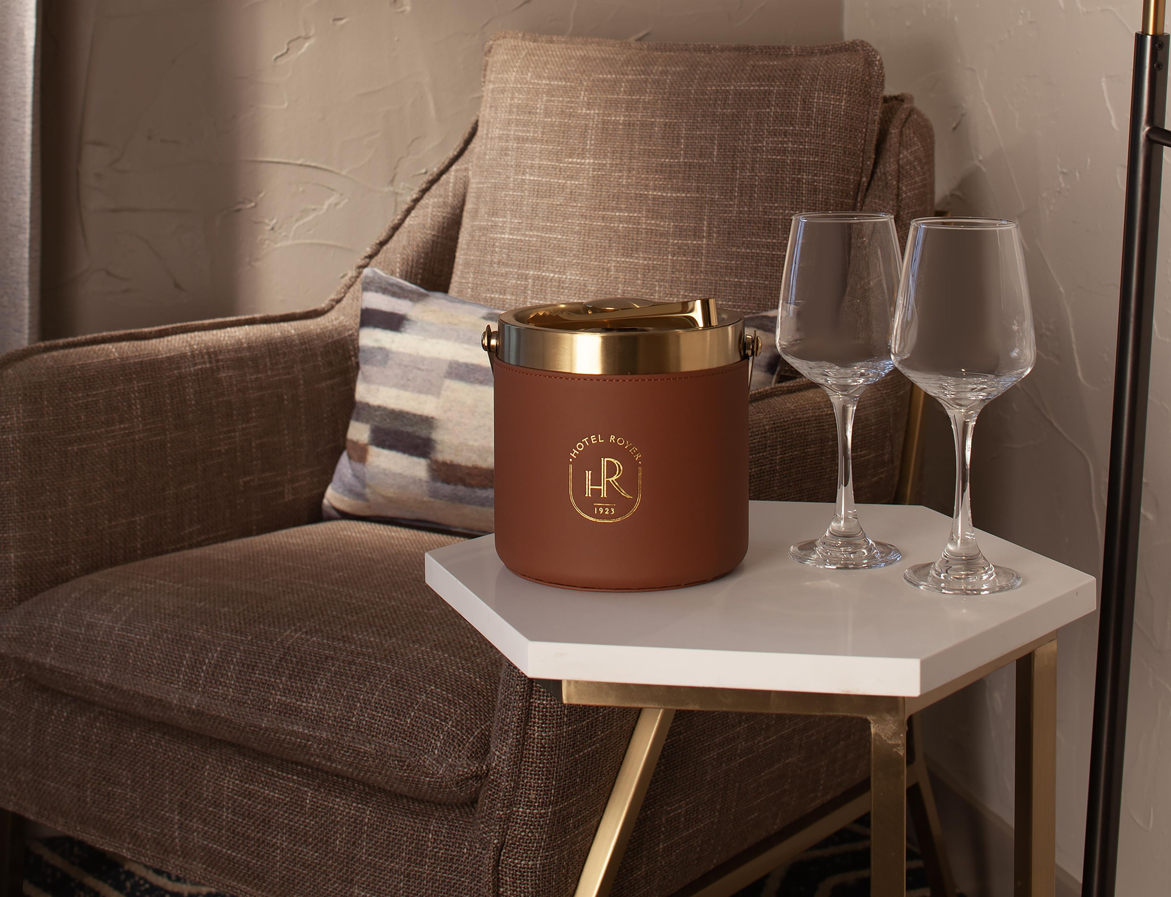Un recipiente de cuero marrón con tapa dorada y las iniciales 'HR' sobre una mesa blanca, junto a una silla tapizada en marrón con un cojín a rayas.