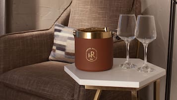 Un recipiente de cuero marrón con tapa dorada y las iniciales 'HR' sobre una mesa blanca, junto a una silla tapizada en marrón con un cojín a rayas.