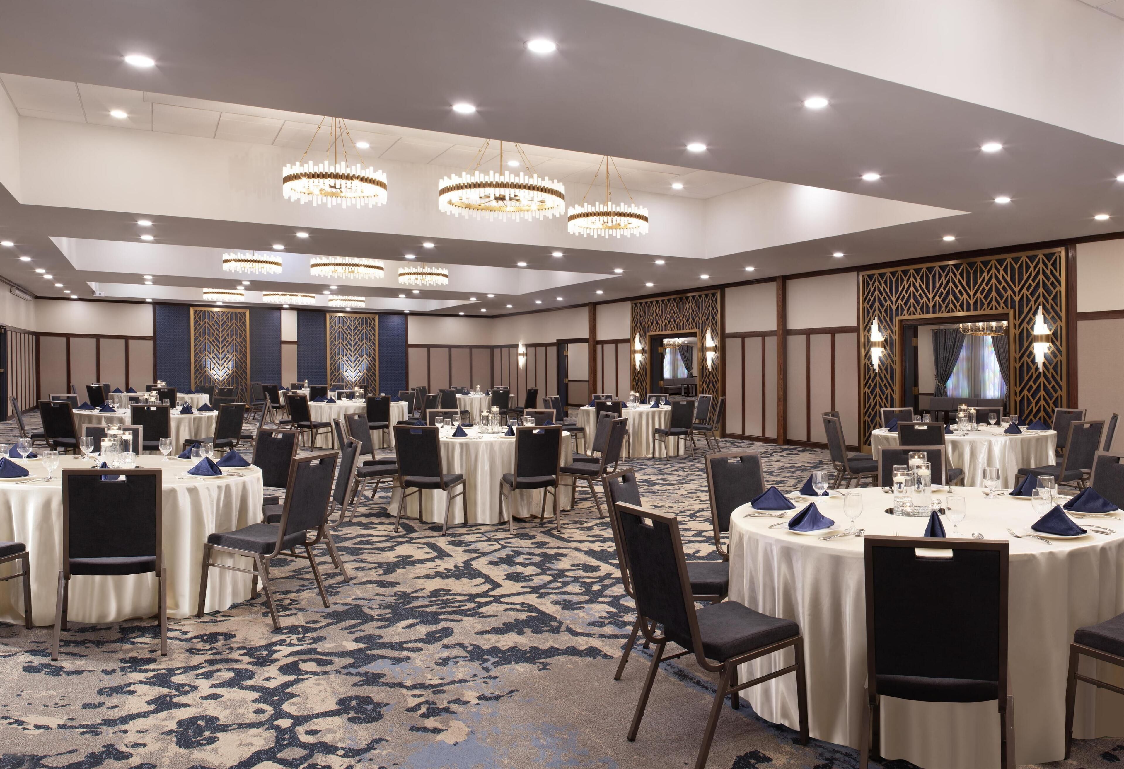 A large banquet hall with round tables, chairs, chandeliers, and a patterned carpet.