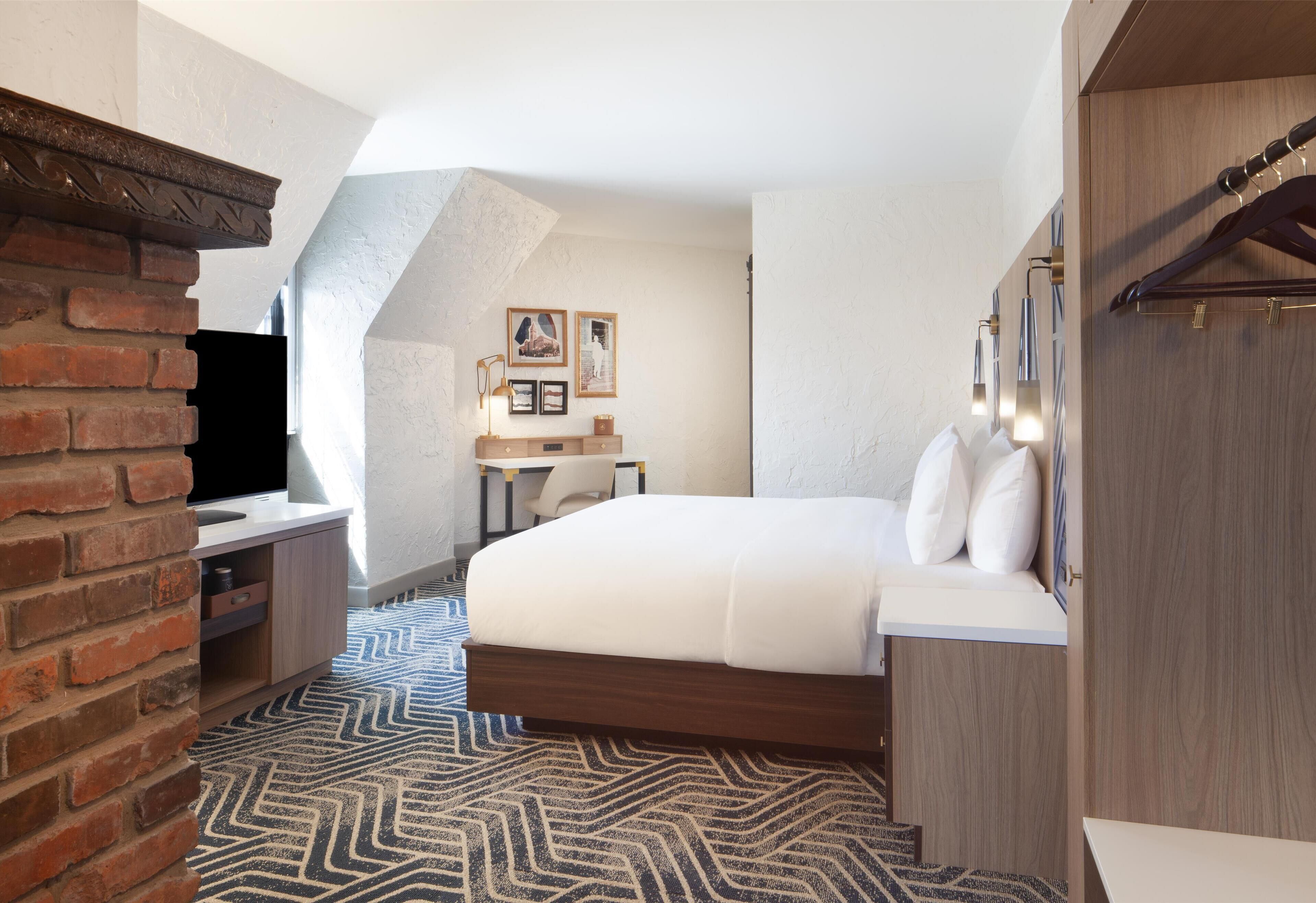 Quarto de hotel com parede de tijolos decorativa, cama com roupa de cama branca, penteadeira de madeira, escrivaninha e uma cadeira, além de tapete estampado.