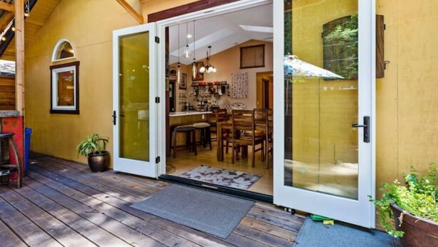 Een houten terras dat leidt naar een huis met een glazen deur, een eethoek binnen en een potplant buiten.