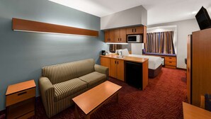 Een compacte hotelkamer met een kitchenette, een sofa, een klein tafeltje en een raam met gordijnen.