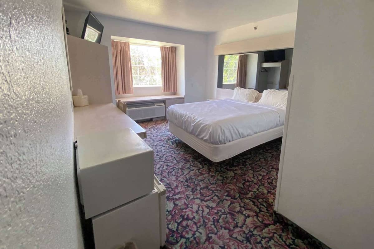 A hotel room with a bed, a desk, a chair, a small refrigerator, and a window with curtains.