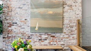 En rustik tegelvägg med en inramad målning av en segelbåt på havet. En träbänk och ett metallbord med en blomsteruppsättning står i förgrunden.