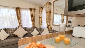 Un salon moderne avec un canapé, une table basse en bois sur laquelle sont posés des croissants, et un coin cuisine équipé d’un grille-pain et de verres à jus.