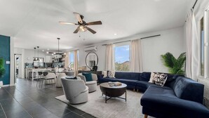 A modern living room with a dark blue sofa, grey armchairs, and a coffee table. There is a ceiling fan, a large window with curtains, and a view of the kitchen in the background. - Desert Artist Oasis| Fire-pit | Spa | 5 min to JT, Desert Artist Oasis| Fire-pit | Spa | 5 min to (Yucca Valley)