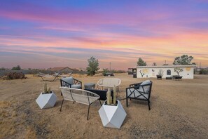 An outdoor seating area with modern furniture and a cactus planter, set against a desert landscape with a building featuring artwork. - Desert Escape w/ Hot Tub + Fire Pit | Near Park (Yucca Valley)
