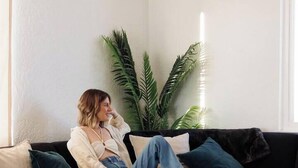 A person sitting on a black sofa in a room with a white wall, a large potted plant, and a round table with a woven basket and candles. - Desert Artist Oasis| Fire-pit | Spa | 5 min to JT, Desert Artist Oasis| Fire-pit | Spa | 5 min to (Yucca Valley)