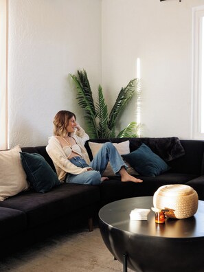 A person sitting on a dark-colored sofa with a round black coffee table in front, a potted plant to the left, and a white wall in the background. - Desert Escape w/ Hot Tub + Fire Pit | Near Park (Yucca Valley)