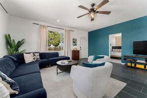 A modern living room with a dark blue sofa, a white armchair, and a coffee table. There is a ceiling fan, a flat-screen TV, and a bookshelf. - Desert Escape w/ Hot Tub + Fire Pit | Near Park (Yucca Valley)